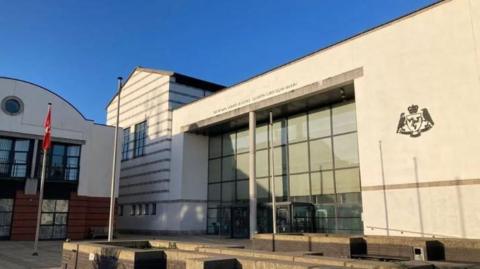 Douglas Courthouse, which is a large white glass-fronted building with the government crest on the wall to the right of the entrance and the words Isle of Man Courts of Justice above it.