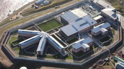 Prison buildings complex with structures of various shapes and sizes seen from above, areas of grass, and an area of sea water nearby.