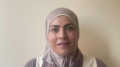Wearing head scarf, striped shirt and white waist coat. Standing in front of a white wall.
