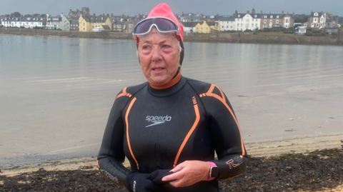 Woman in a Speedo wetsuit, large goggles and a pink swim hat. She is standing on a sandy beach, at the water's edge. Houses a visible on the other side of the water. 