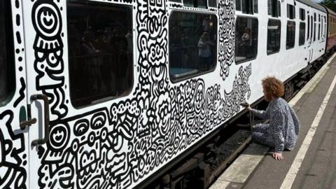 Mr Doodle - a man with ginger hair and a doodle costume on - doodling on the side of a train carriage while sitting on a platform