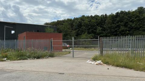A small brick building is behind a barbed wire gate and metal fence. There is rubbish blown against long grass near the entrance.