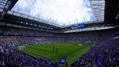 View from an interior corner of the new stadium showing fans in blue packed in all four stands around the pitch. Both teams walk out from the tunnel on the right before the match. Blue club flags are flying across both stands on left and right. Blue flares or fireworks have been let off from the roof overhead with white clouds above in the blue sky.