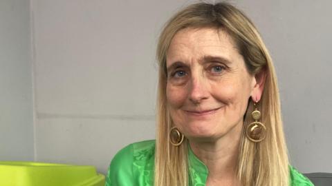 A woman with blonde hair and a green shirt with large gold hoop earrings. She is looking into the camera and smiling.