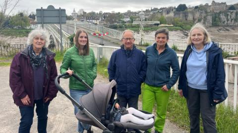 Image shows a group of people stand in a car park in front of a bridge, which is blocked by two barriers. The group consists of three women and two men, one of whom is with a pushchair. In the background, a river flows beneath the bridge, and beyond it stands an old castle. 