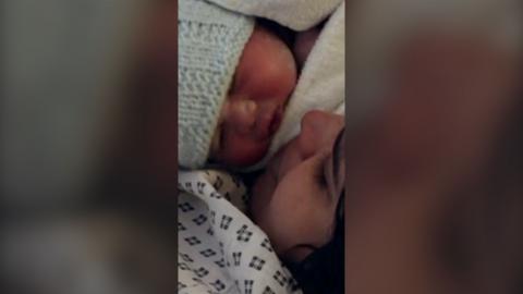 A mother with dark hair in a hospital gown, looking into the face of her baby just born by C-section.