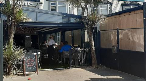 Street view of the restaurant showing a glass extension between the pavement and the restaurant. People are sitting in the extension while a waitress in a white shirt and with blond hair in a ponytail serves them. There is an A-board outside that says 'please wait to be seated'