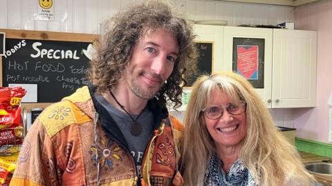 Chris Carrier and Jacs Guyton pose together in a cafe. Mr Carrier has a beard and is wearing a flowery top and Ms Guyton has long blonde hair and is wearing glasses. They are both wearing brightly printed clothes. 