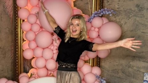 Victoria Skinner, with her arms outstretched, smiling, with her mouth open, in front of lots of pink balloons. She is wearing a black top, with a necklace, and a gold skirt, she is by a large mirror in a room. 