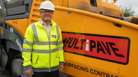 Councillor Rupert Swarbrick is smiling as she stands next to a roadworks vehicle. He is wearing a hi-vis jacket with the Lancashire County Council logo on it, along with a safety helmet.