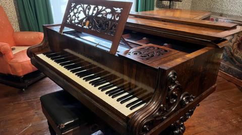 Image of a small ornate grand piano with a leather piano stool.  The piano is in a period room.  In the background is a pink damask fireside chair, a window with green curtains and a tapestry on a wall.  The walls are wallpapered with green floral wallpaper and the floor is wood.
