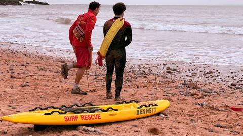 Two RNLI lifeguards with their backs to the camera stand by a yellow surfboard on a beach one wears red jacket and shorts while the other wears a black wetsuit
