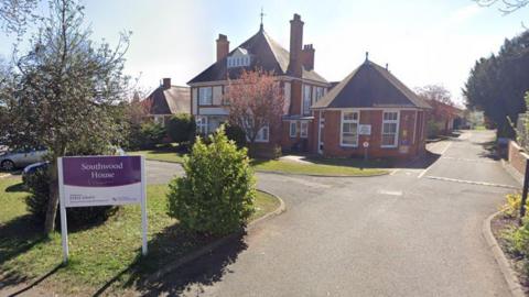 A red brick care home in the background. A grey driveway leads up to it and and there is a white sign in the foreground which states Southwood House. 