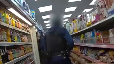 The back of a Met Police office raiding a shop. There are drinks in a fridge to the left and food items on shelving on the right.