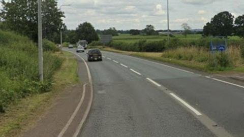 A road, with numerous cars travelling in both directions, is seen curving to the left. There are hedges and fields in the distance and a footpath in the foreground.