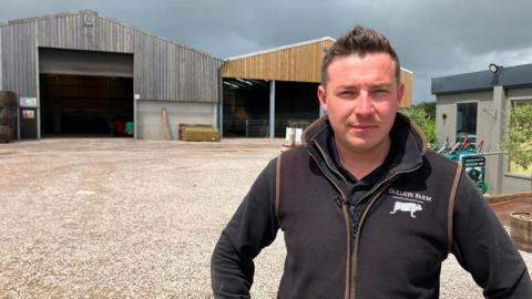 A man stands outside farm buildings, with barns and equipment in the background.
