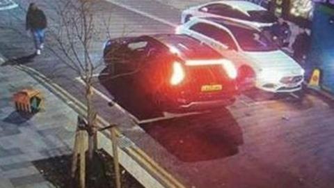 A low resolution CCTV image of two cars in the road, one black and one white, facing in opposite directions. The rear lights of the black care and headlights of the white car are on. There are two people standing by the side of the white car. 