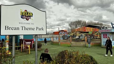 A large notice in the foreground saying 'Welcome to Brean Theme Park' and fairground rides in the background on a grass pitch with a few people milling around. The clouds are grey and gloomy