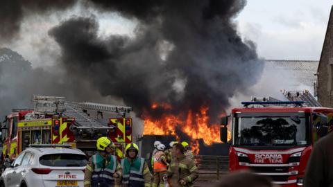 Firefighters respond to a large blaze at a farmyard, with flames and thick black smoke billowing high into the sky. Several fire engines and emergency personnel are positioned around the burning structure, while nearby buildings and vehicles are partially obscured by smoke.
