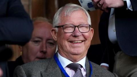 A man with white hair and glasses, wearing a grey suit with blue lanyard, sitting in the stands at a football match.