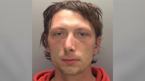 A police mugshot of a man with short dark brown hair wearing an orange sweatshirt. He is looking straight at the camera.