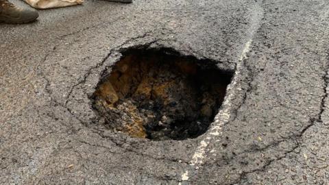 A hole in concrete. Some chunks of concrete have fallen into the floor. It is unclear how deep it is due to the angle of the picture. There are lines surrounding the hole where the ground has cracked further.
