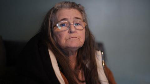 An elderly lady stares at the camera with a neutral expression. She has dark brown hair, gold-rimmed glasses and is wearing what appears to be a brown jacket with a white lining.