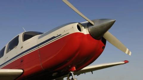 Single-prop aircraft with red bottom half and white top half. There are windows around the cockpit. The propeller is grey and has a dark grey nose cone.  