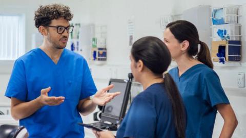 One male and two female nurses in blue scrubs stand in a hospital room.