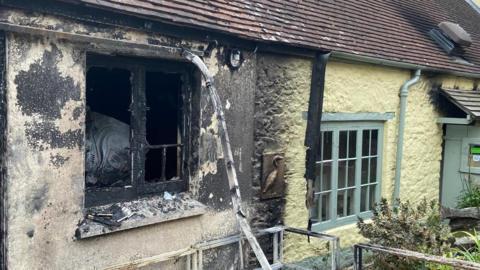 The back of a house is covered in black ash. The window is broken and burnt. The house next to it looks clean and has yellow paint.