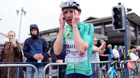 A man wearing a cycle helmet and a turquoise cycling outfit holds his head in his hands as he wins the Tour of Britain. Crowds behind him and barriers line the street