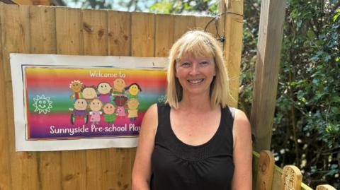 A lady in a black vest top stands in front of a wooden fence with the sign to a preschool attached to it.