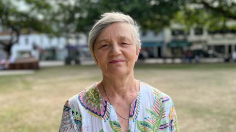 Deputy Mary Le Hegarat, Minister for Justice and Home Affairs. She has short dark grey hair and is wearing red lipstick. She is wearing a white blouse with colourful flowers on it.