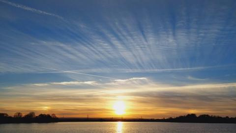 On the horizon, above a wide area of water, the sun is a golden ball setting in a horizontal layer of yellow sky. Above wisps of white cloud stretch vertically in a blue sky