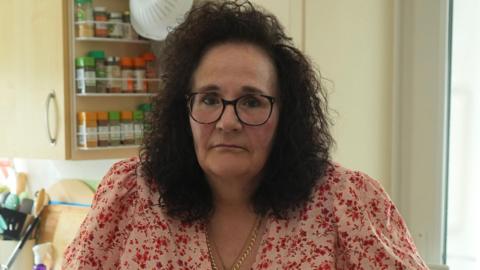 Ms Stocker is on crutches and is standing in her kitchen. She is wearing a red summer dress with sleeves, gold necklace and glasses. She has medium length brown curly hair and is not smiling. 