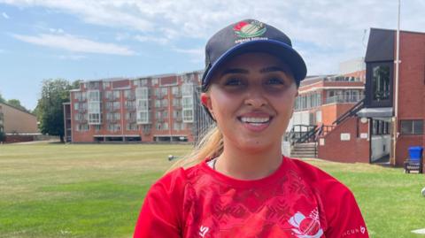 Firooza Amiri smiles at the camera while standing next to the cricket pitch. She wears the red T-Shirt of her kit, and a black baseball cap. Her fair hair is tied back. 