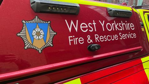 The side of a bright red emergency response vehicle belonging to the West Yorkshire Fire & Rescue Service. The service’s name is printed prominently in large white letters on the vehicle’s body. To the left of the text is the organisation’s crest, which features a white rose at its centre set against a blue and gold shield, all framed by a stylised star-shaped emblem.