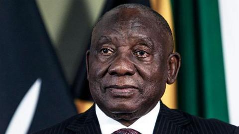 A head-and-shoulders image of Cyril Ramaphosa. He is wearing a dark suit and the white collar of his shirt can just be seen. Some flags are out of focus in the background.