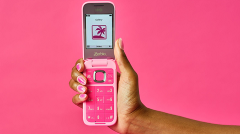 A hand holding the phone. It is a bright pink early 00s style flip phone, with big numbers and a small screen. The person has pink nail polish.