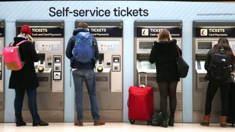 Commuters with backpacks and luggage buy tickets at self-service machines in London Waterloo station