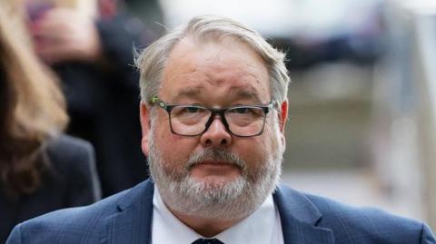 Close up of Christopher Mills outside court. He wears a blue suit, a white shirt and has black glasses. He has short grey hair and a grey beard. 