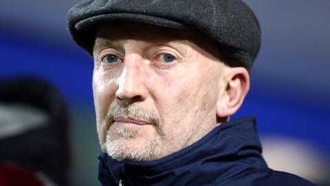 A close-up of Ian Holloway, showing his head and neck. He is wearing a navy coat with a high neck zipped all the way up and a dark grey baker boy-style hat. He has a short, grey moustache and goatee. 