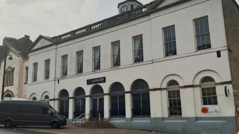 The outside of Quayside Exchange which is a large two-storey white building with a clock tower on its roof. The bottom floor windows are large and arched, which the upper floor windows are rectangular. There is a black sign above the door which reads 'Lasgidi' and a black van parked at an angle in front of it. The path in front of the building is sloped from left to right and the sky above is grey.