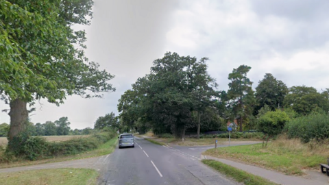 A silver car drives away from along a road, bordered by grass verges, trees and fields. There is a crossroads.