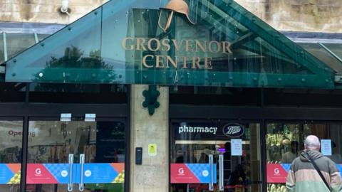 A glass frontage with eight double glass doors and a green glass rain cover displaying the sign for the Grosvenor Centre.