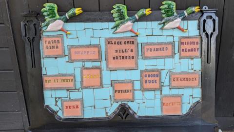 Ceramic artwork with three flying ducks at the top of a frame. Turquoise ceramic tiles are interspersed with other larger tiles with phrases or words on including tater dina, snappin, lobby, puthery, channock and buzz