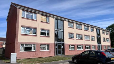 Picture shows a three storey block of flats, with cars parked in front. An upstairs  window is boarded up. 