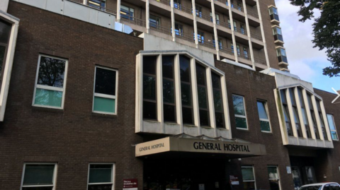 A photo of the front entrance of Jersey General Hospital. It is a large brown brick building.