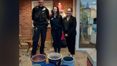 A male police officer stands next to two women behind some plant pots