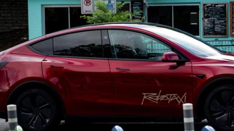 A red Tesla vehicle with a "Robotaxi" logo on the side of it is shown on a street in Austin, Texas. A human operator can be seen in the car's passenger seat.
